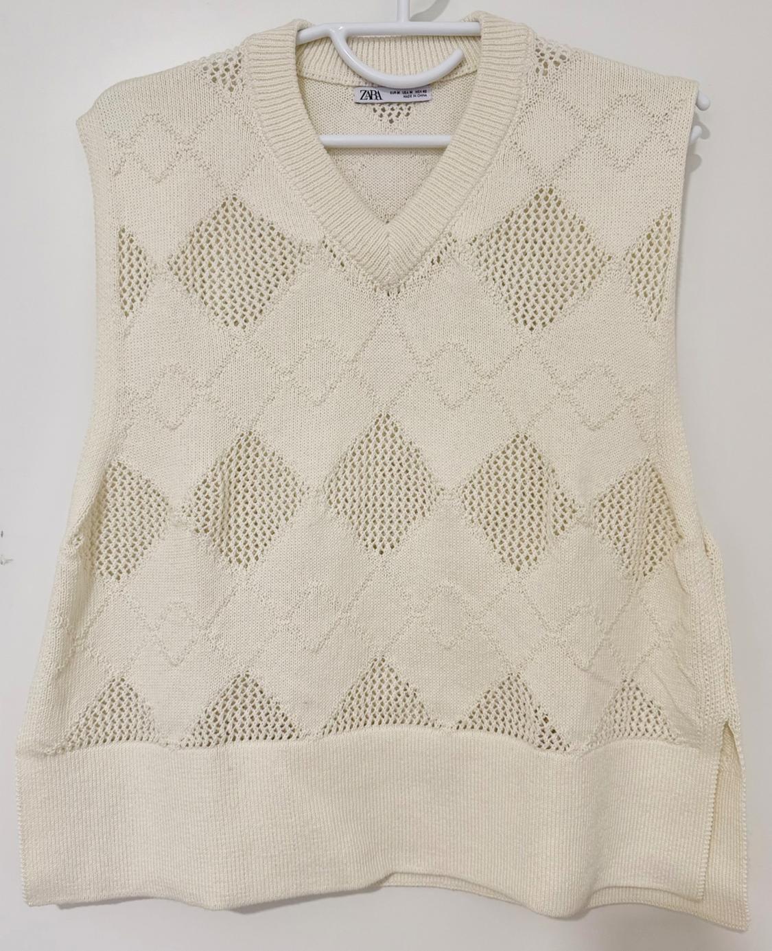 Beige knitted waistcoat with openwork diamond design, hanging on a white hanger. Label visible on the collar.
