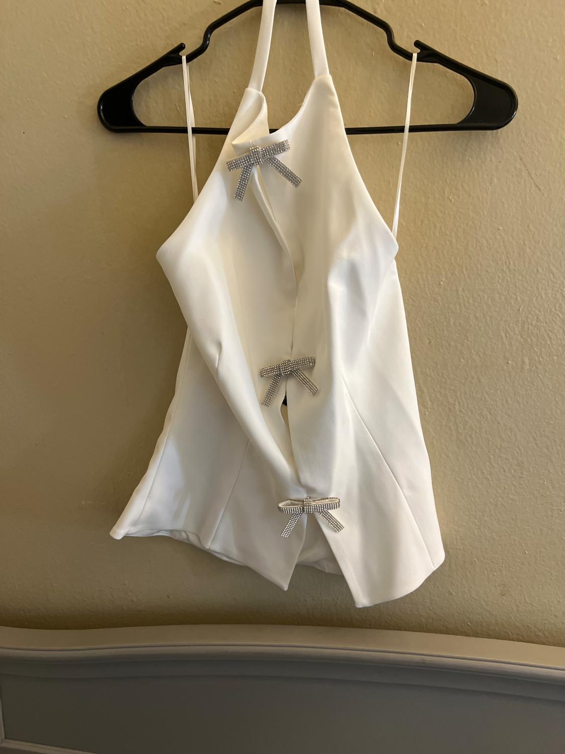 White tank top with thin straps and three shiny decorative bows, hanging on a black hanger against a beige wall.