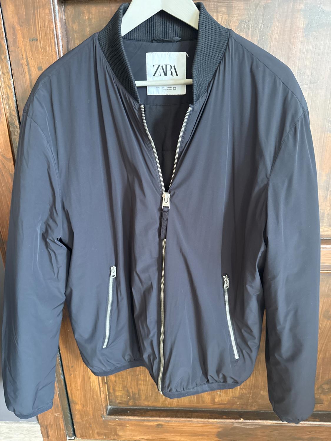 Black jacket with a zip and pockets, hanging on a hanger in front of a wooden door. Visible label: ZARA.