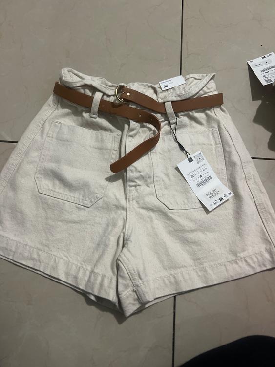 Beige shorts with a brown belt on a tiled floor. Visible tags with size 38 and barcode.