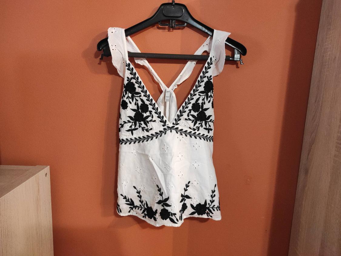 White blouse with black floral embroidery hanging on a black hanger against an orange wall.