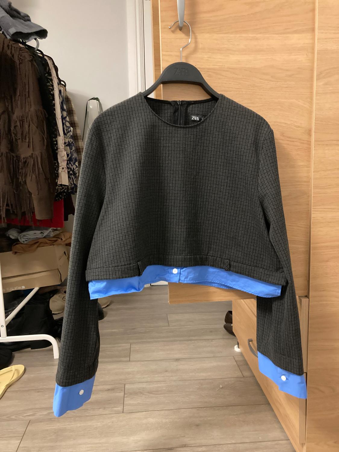 Grey jumper with blue cuffs and hem, hanging on a black hanger in a wooden wardrobe. Background with hanging clothes.