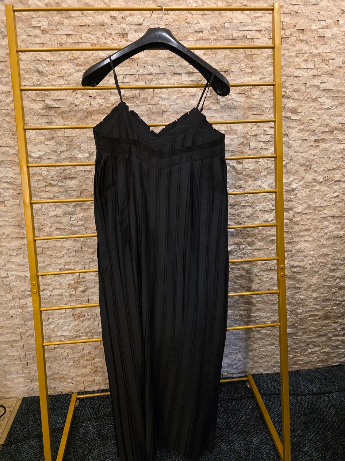 Black spaghetti strap dress with vertical pleats, hanging on a black hanger over a gold frame and brick wall.