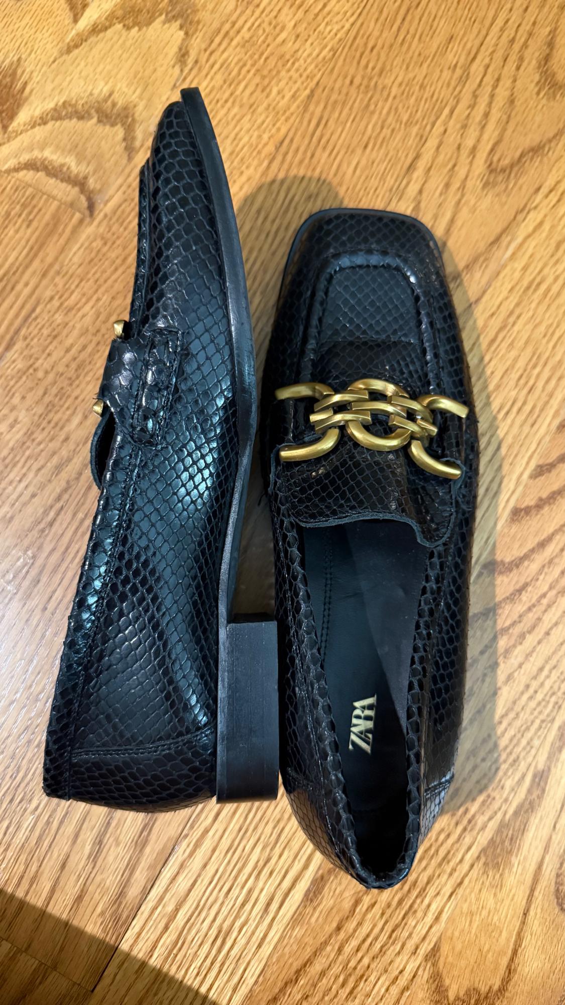 Black shoes with snakeskin texture and gold chain detail on the upper, on a wooden floor. Visible text: ZARA.