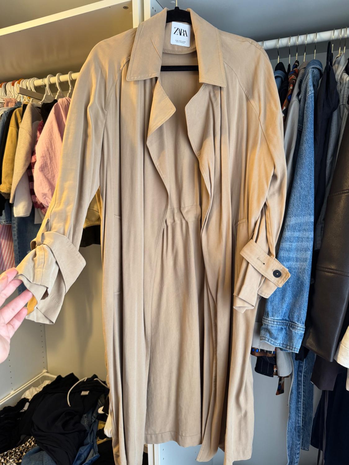 A beige trench coat hangs inside a closet next to other pieces of clothing. A hand brushes against its sleeve. The label shows the name ZARA.
