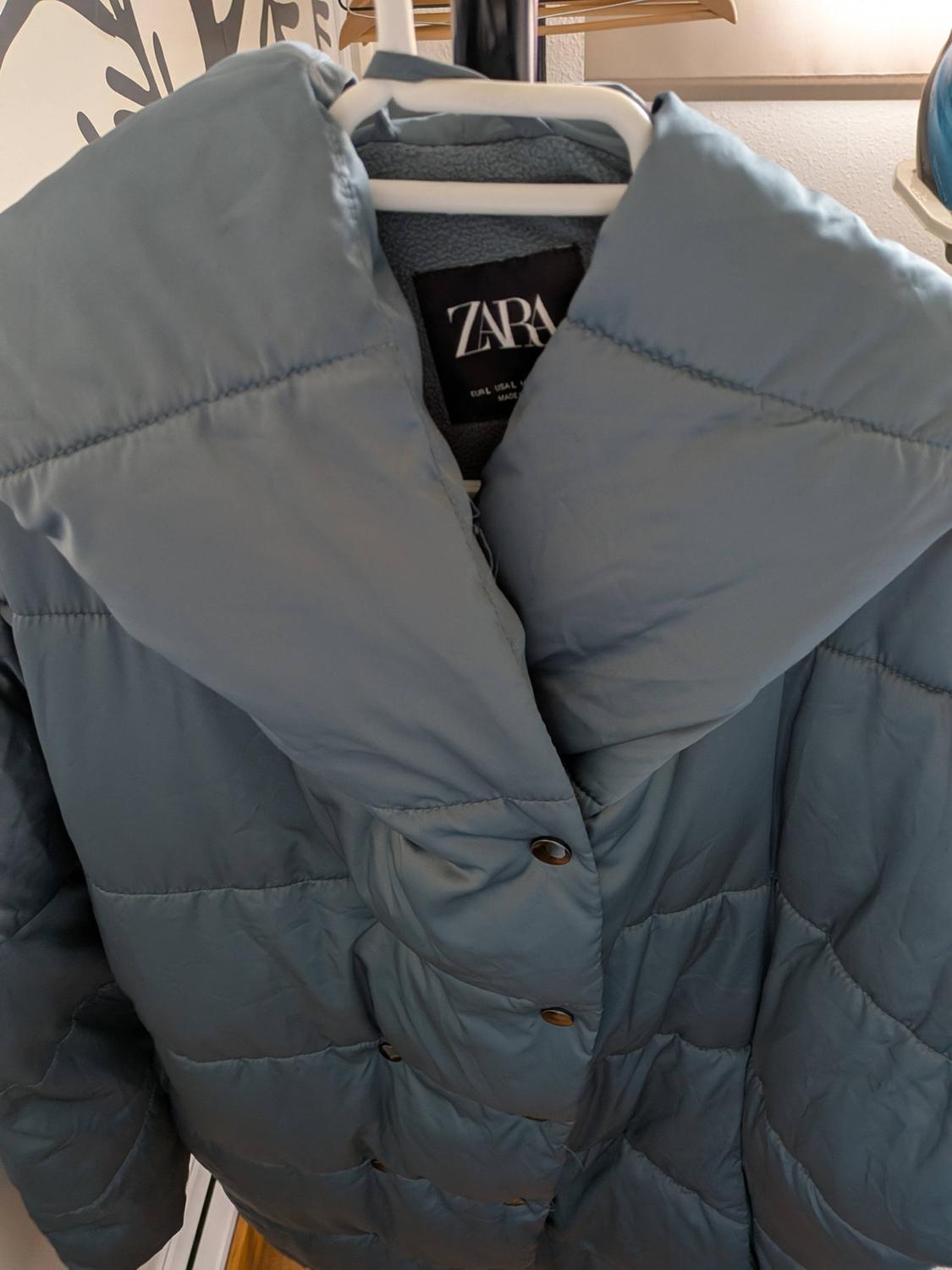 Blue padded coat hanging on a white hanger. Visible label with the text "ZARA". Abstract design background on the wall.