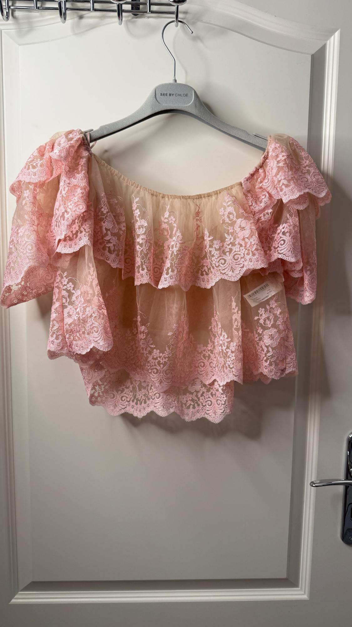 Sheer pink top with lace ruffles hanging on a grey hanger with the text SEE BY CHLOÉ, against a white door.