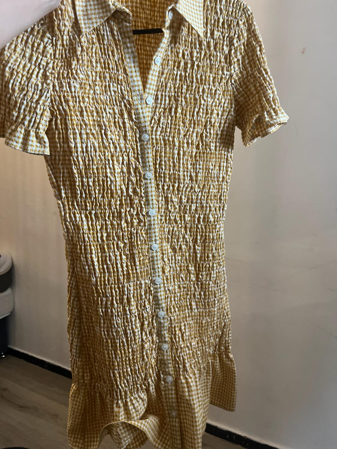 Yellow and white gingham shirt dress with a gathered texture, short sleeves and front buttons, held up in front of a wall.