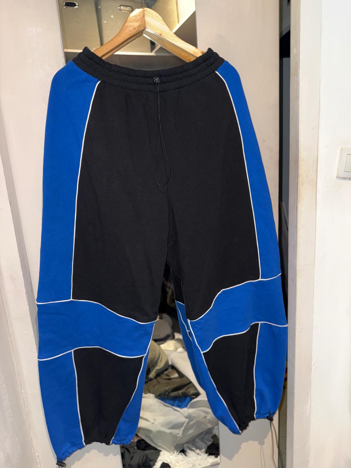 Sports trousers hanging on a hanger, with a black and blue colour-block design and white line detailing. Indoor background.