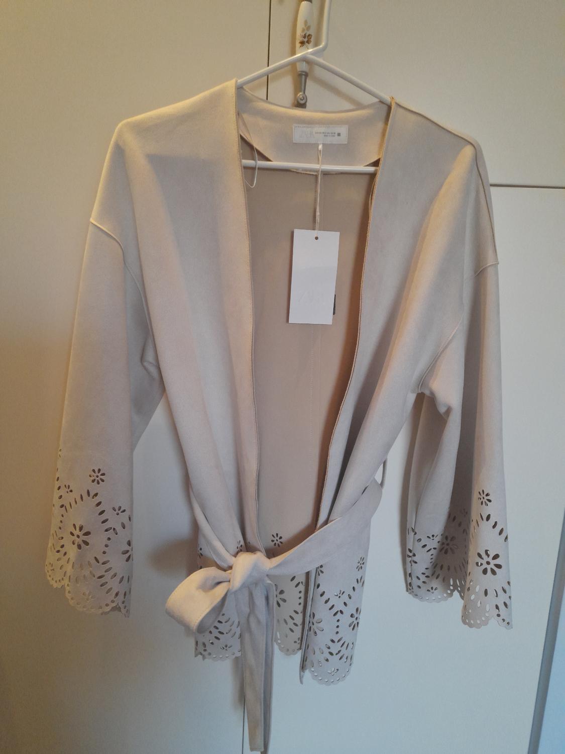 Beige jacket hanging on a hanger, with openwork details on the sleeves and bottom hem. White tag hanging from the collar.