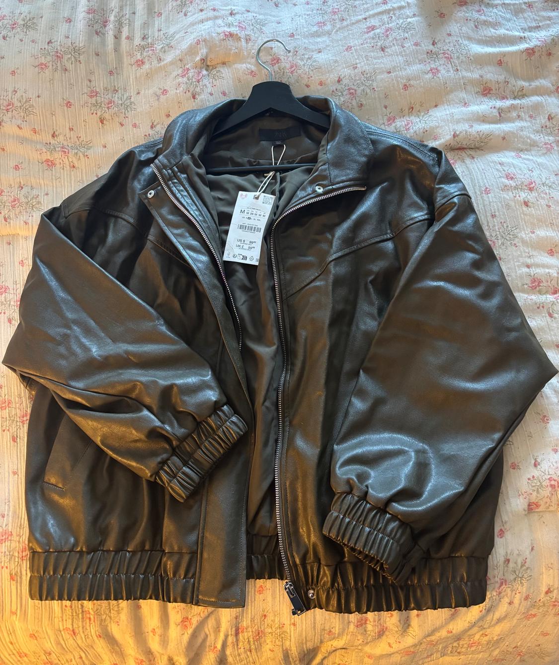 Black synthetic zip-up jacket, hanging on a hanger over a floral quilt. Visible tag with product details.