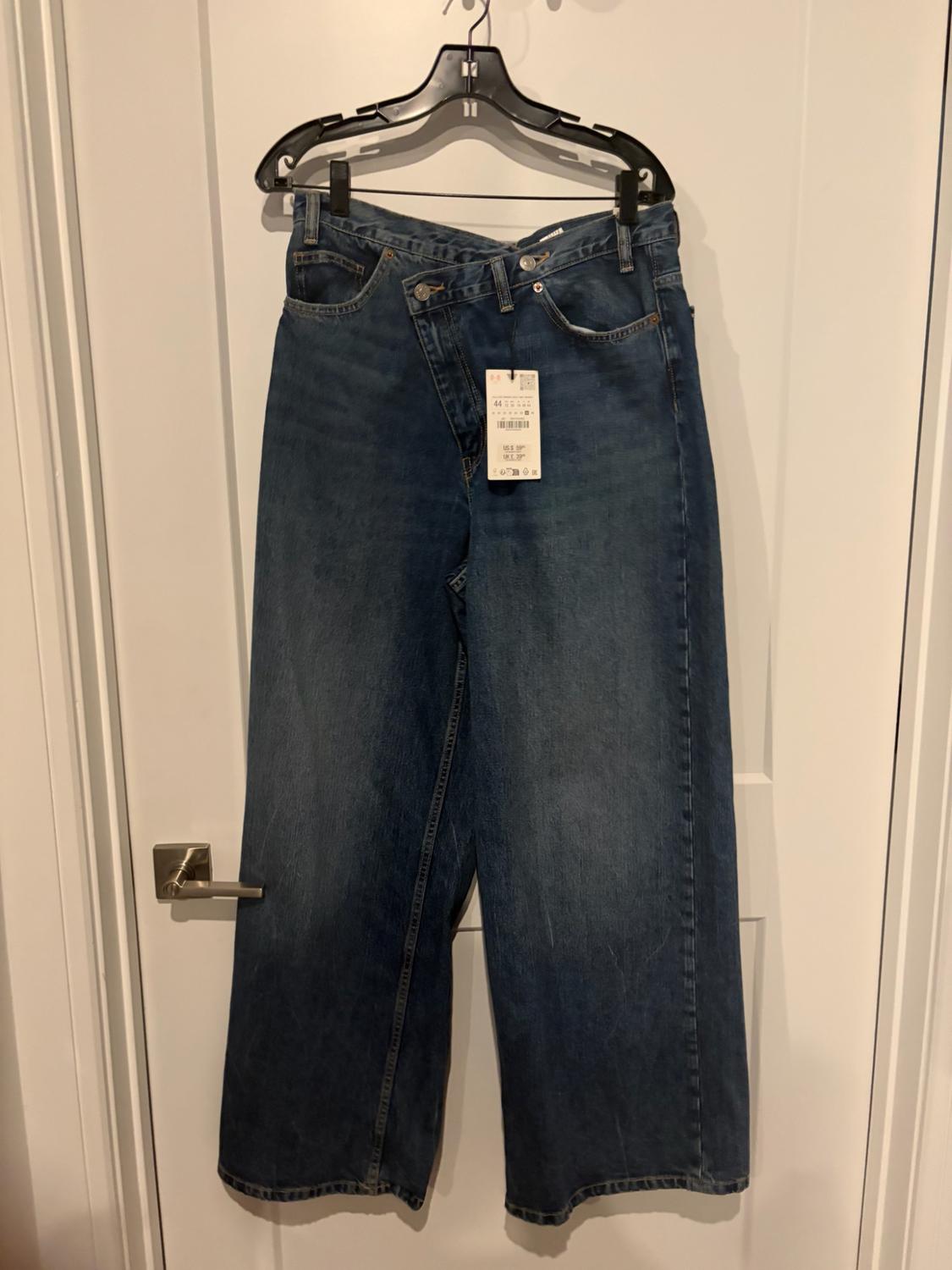 Wide-leg blue jeans with an asymmetric crossover waist and tag, hanging on a black hanger on a white door.