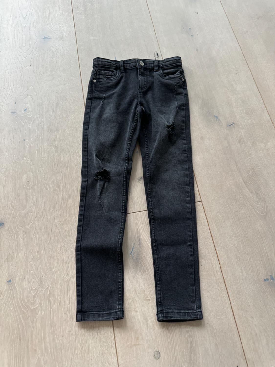 Distressed black denim jeans with rips on the legs, placed on a light wooden floor.