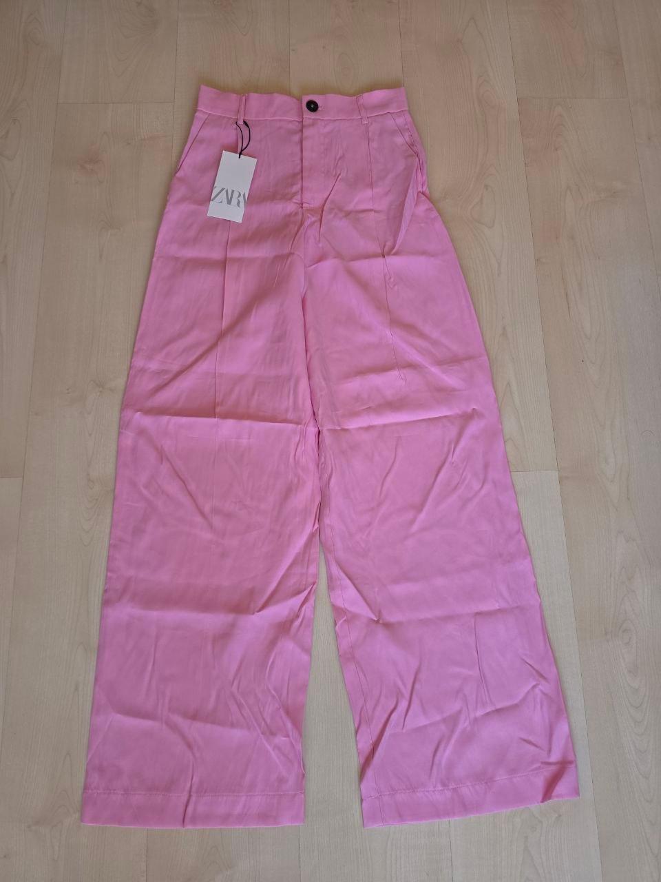 Pink wide-leg trousers with a swing tag that reads "ZVR", placed on a light wooden floor.