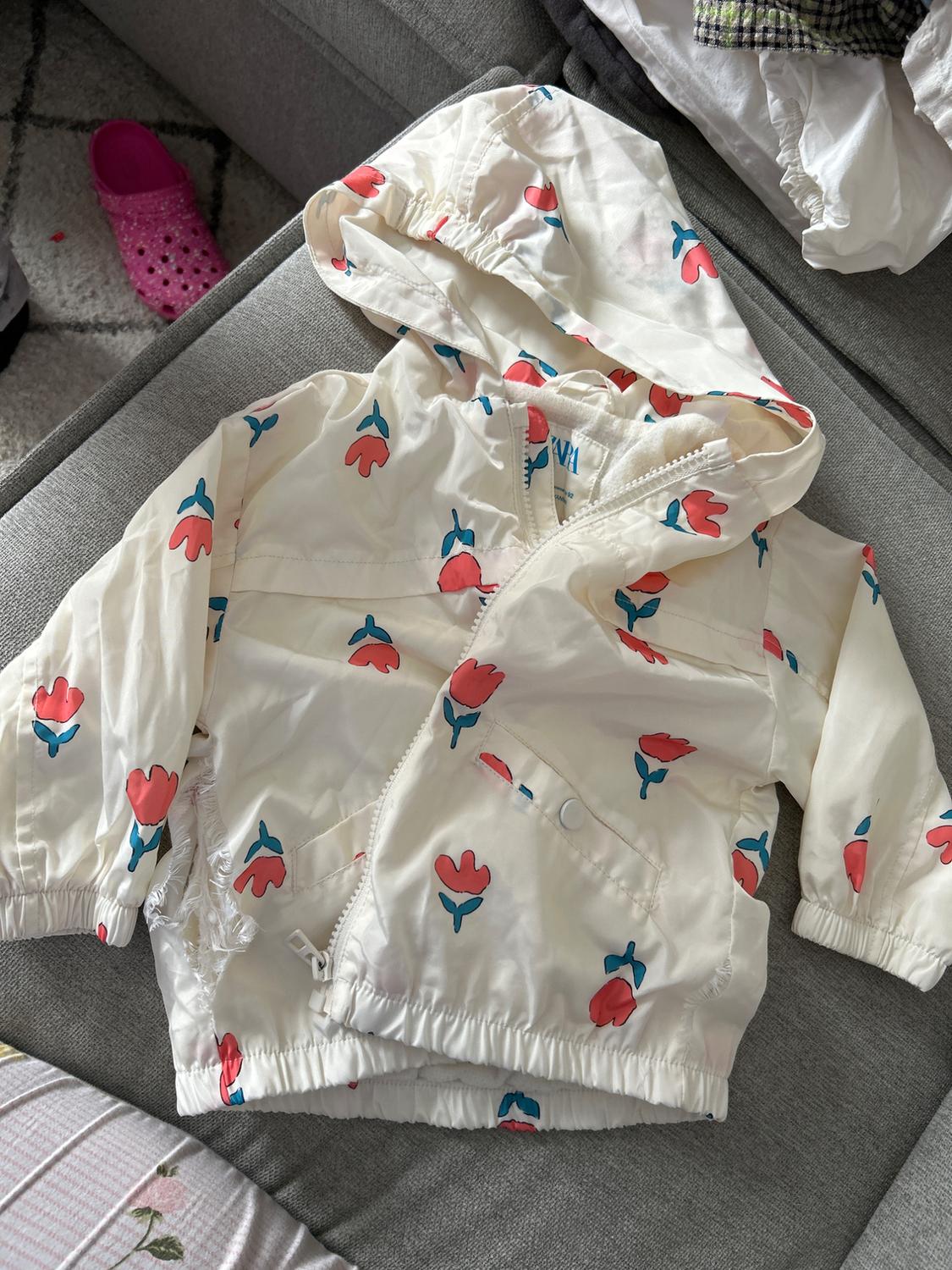 White children's jacket with red and blue floral print, hood, and front fastening, on a grey sofa.