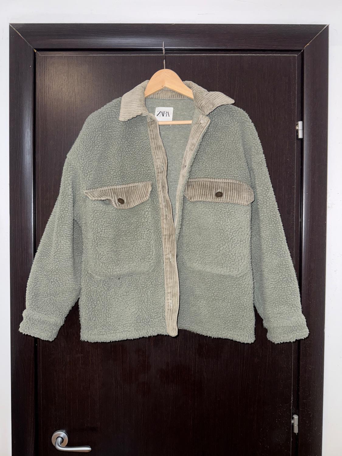 Green wool jacket hanging on a dark door. Corduroy details on the collar and pockets. Visible label with text "ZVΛ".