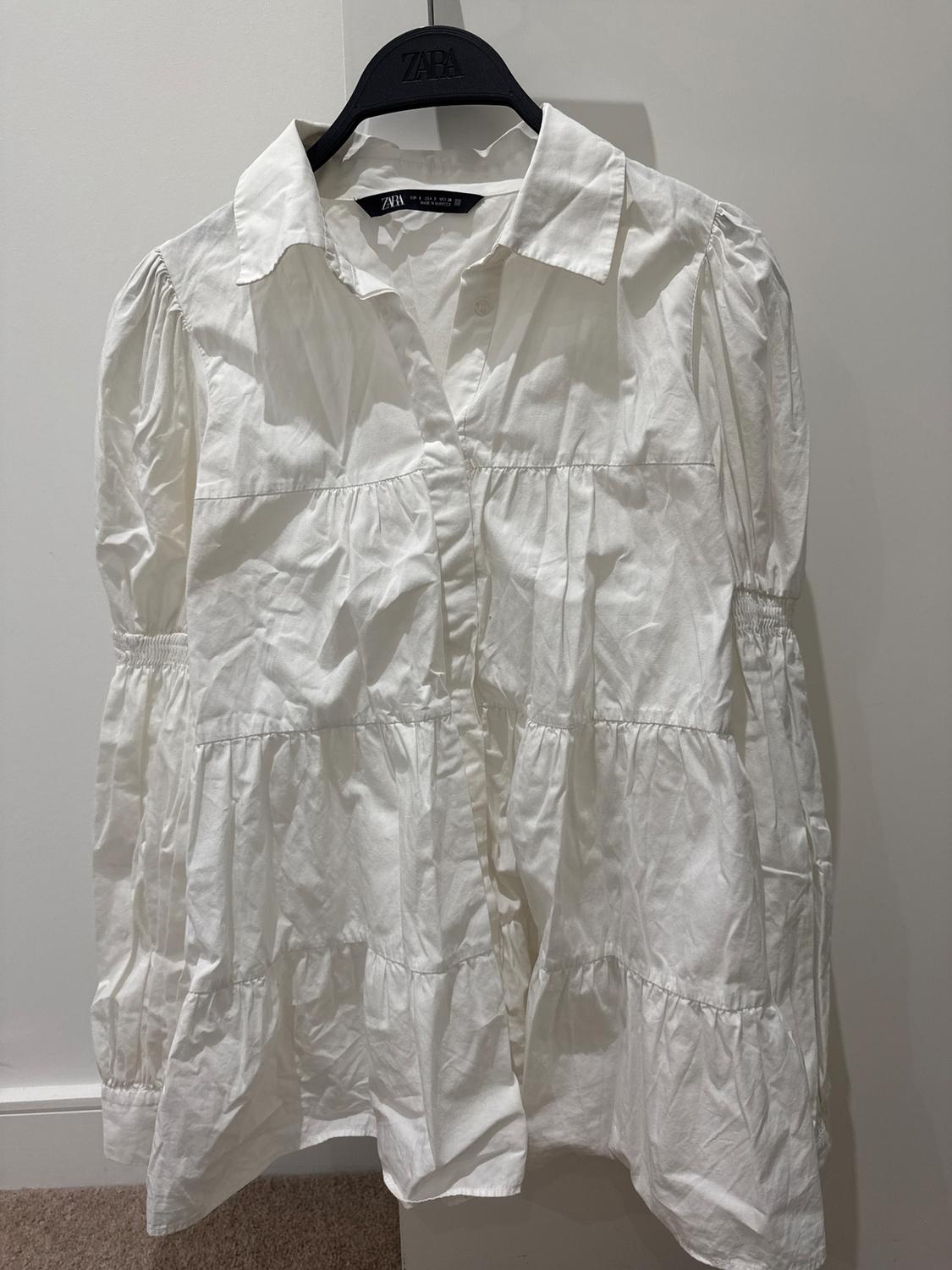 White long-sleeved blouse with ruffles, hanging on a black hanger with the brand visible. White background and beige floor.