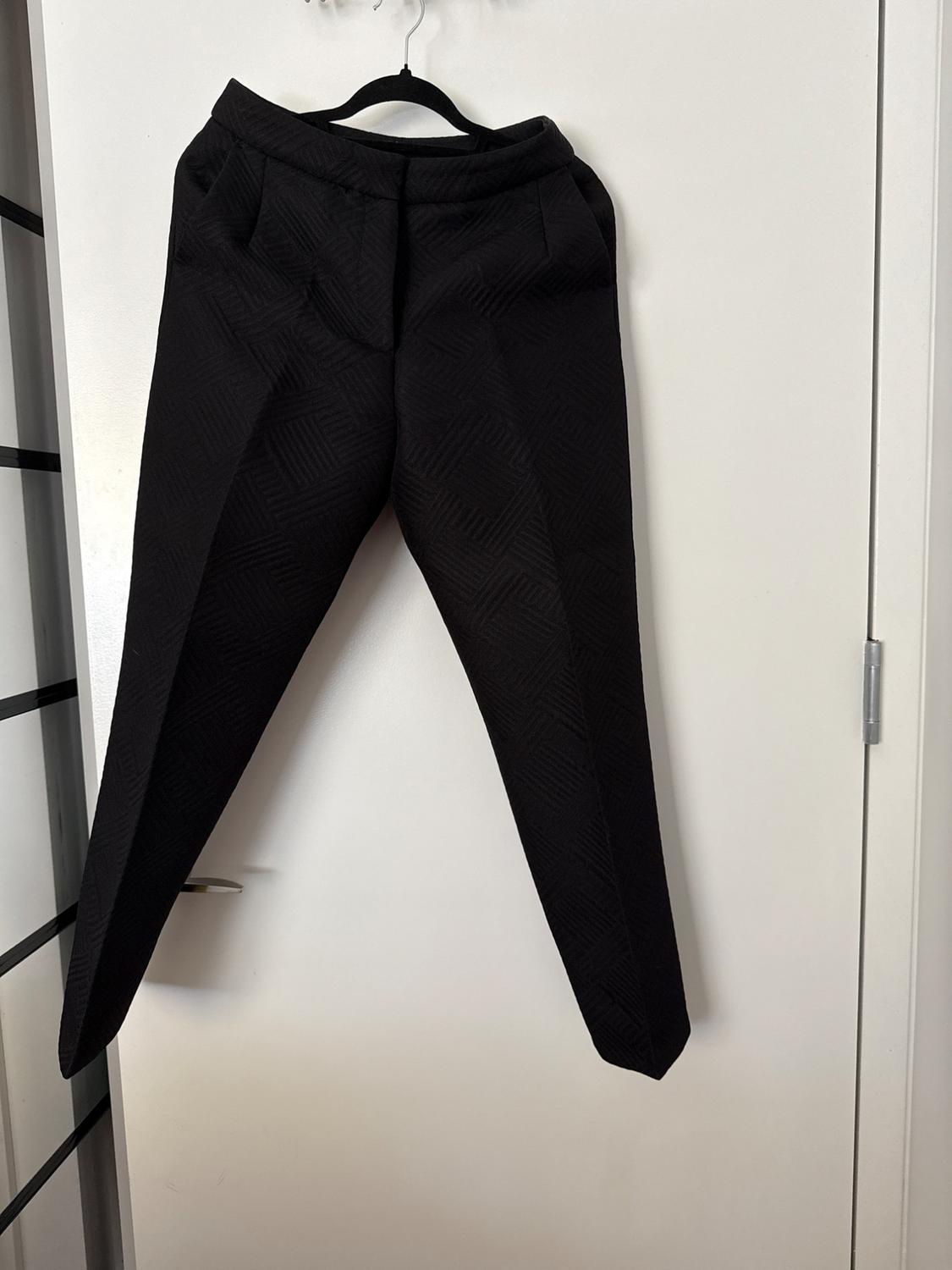 Black trousers with a geometric pattern hanging on a hanger on a white door with a silver handle. Casual setting.