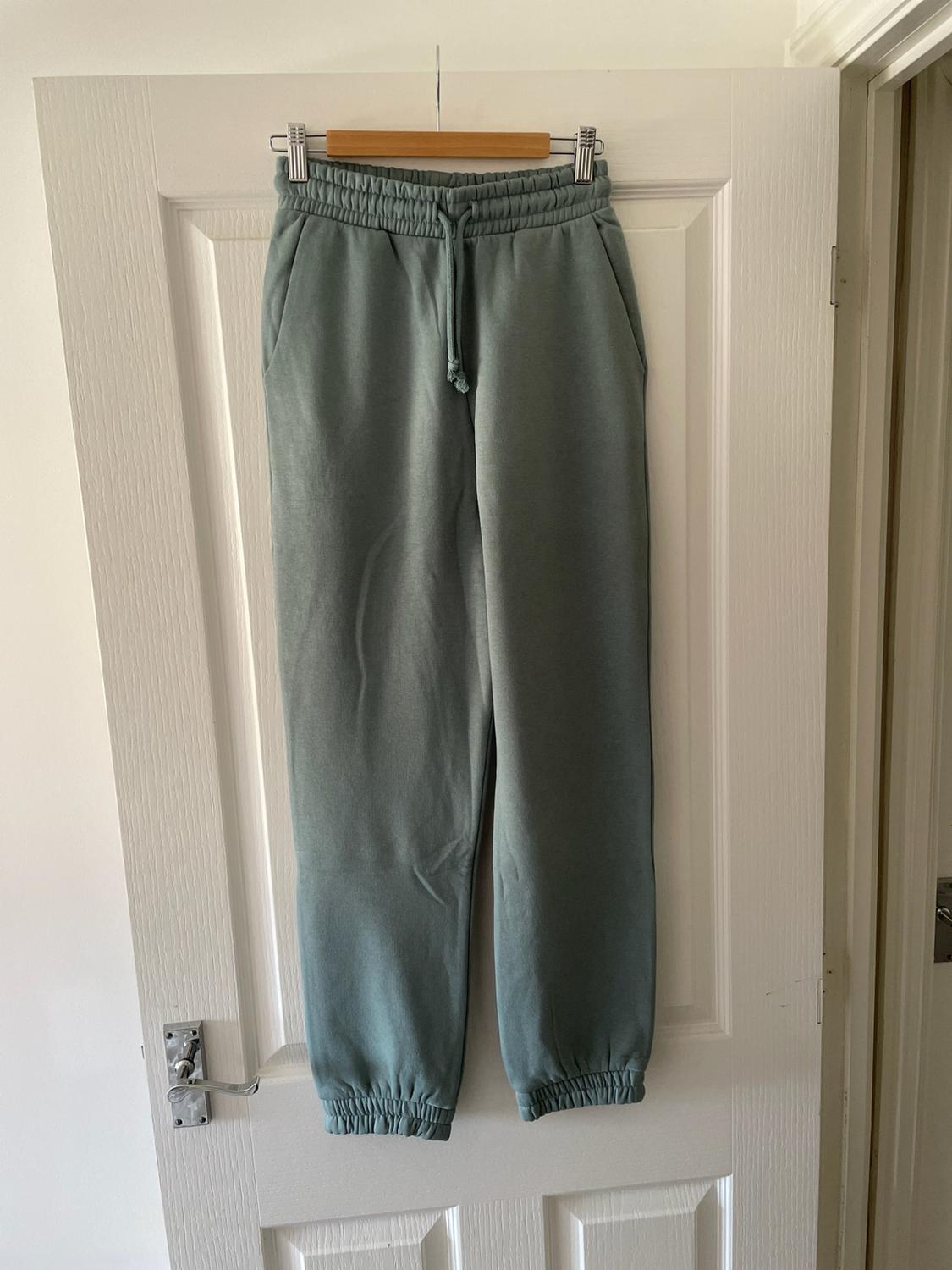 Light green tracksuit bottoms hanging on a wooden hanger on a white door.
Elasticated waist with drawstring and cuffed hems.