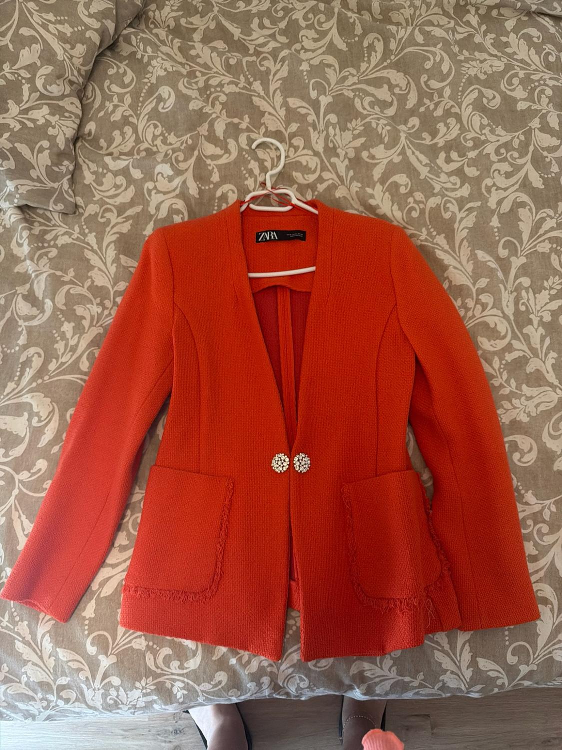 Textured orange jacket with a ZARA label on a hanger, jewelled buttons and frayed pockets, on a bed with a floral print.