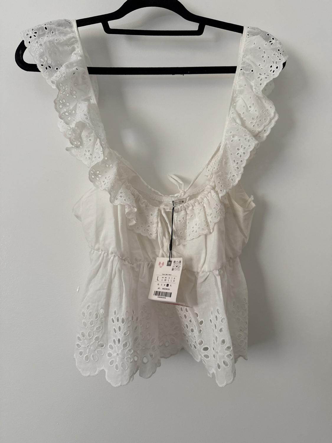 Relaxed-fit white top with ruffles and broderie anglaise detailing, on a black hanger against a white background. Label visible with the letter L.