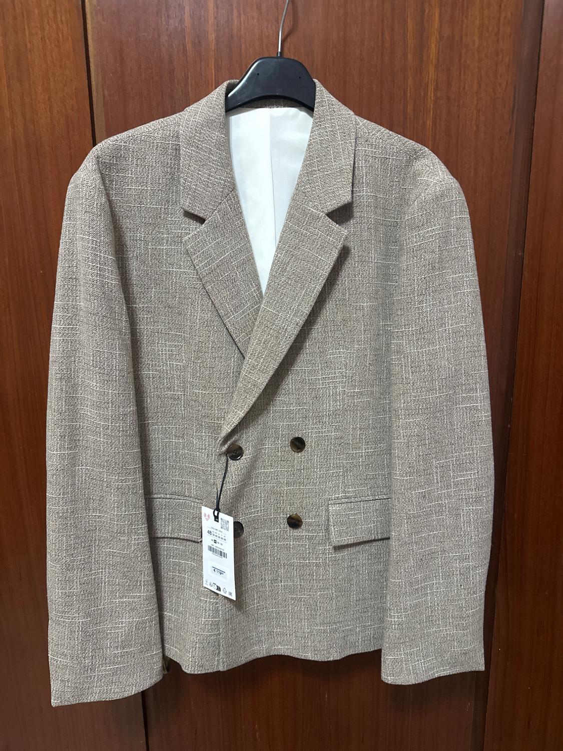 Beige double-breasted jacket hanging on a black hanger, with visible tag, against a brown wooden background.