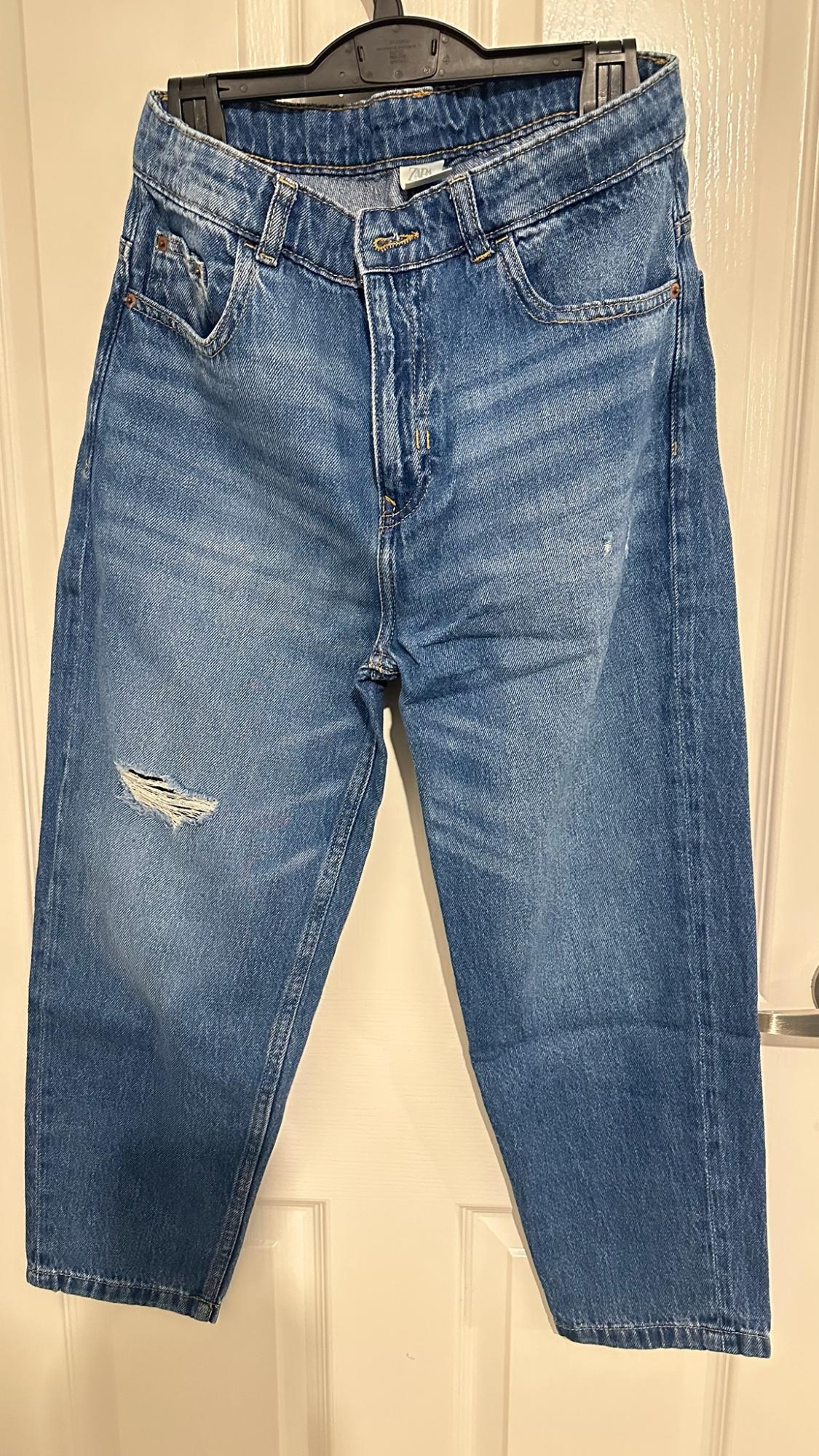 Blue jeans with a ripped leg, hanging on a black hanger over a white door. ZARA is written on the inner label.