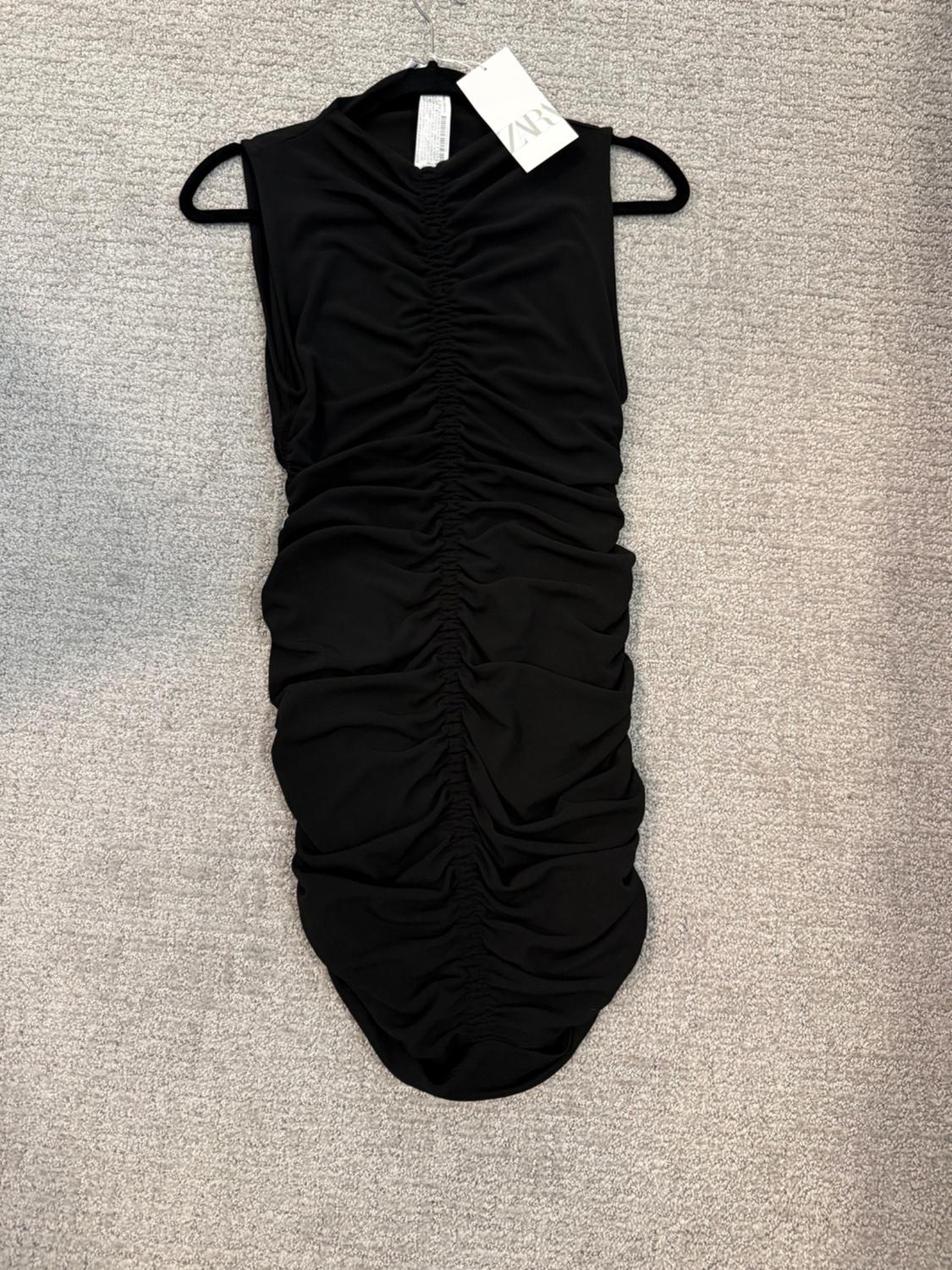 Sleeveless black dress, high neck with central ruching, on a black hanger on a light gray carpet. Tag with ZARA text.