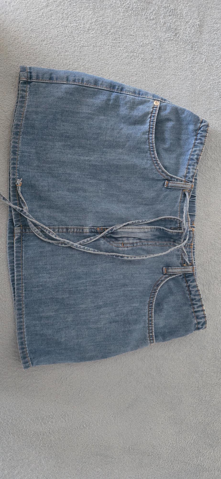 Light blue denim skirt with tie belt, front pockets and visible stitching, on a light grey background.