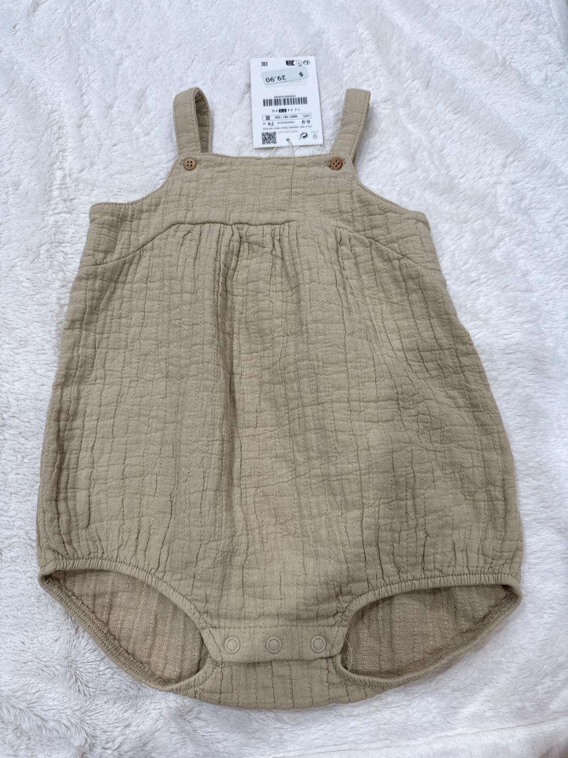 Beige baby romper with straps and wooden buttons, on a white background. Visible label with a price of 29.90.