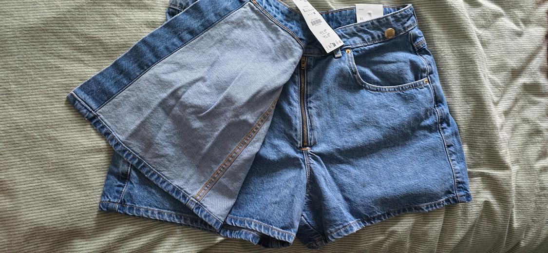 Blue denim shorts with zip and gold button, on striped fabric. Visible labels with size and price information.