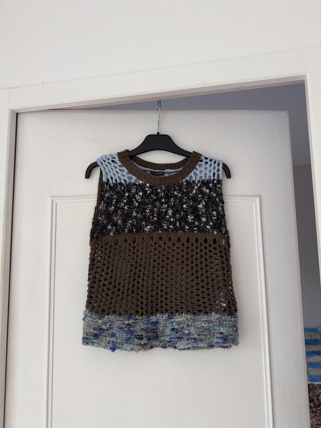 Multicoloured knitted waistcoat hanging on a white door. Mesh design in shades of brown, black, blue, and grey.