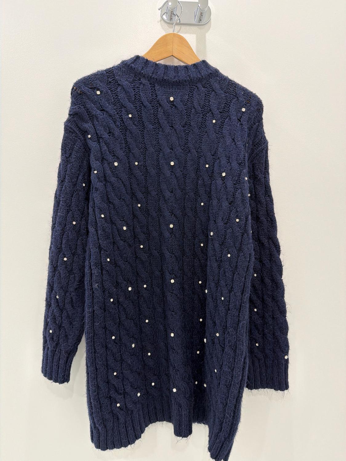 Navy blue cable-knit sweater with sparkly appliqués, hanging on a wooden hanger against a white wall.