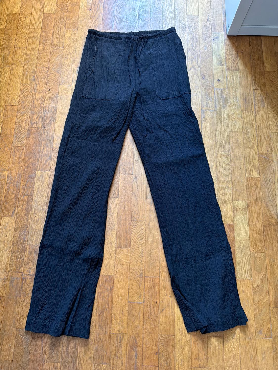 Black textured-fabric trousers with an elasticated waistband, drawstring and front pockets, laid out on a wooden floor.