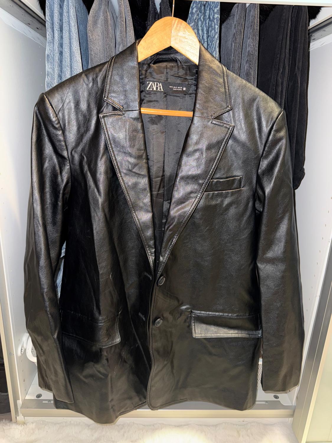 Black leather jacket hanging on a wooden hanger, with a visible Zara label, in a wardrobe with clothes in the background.