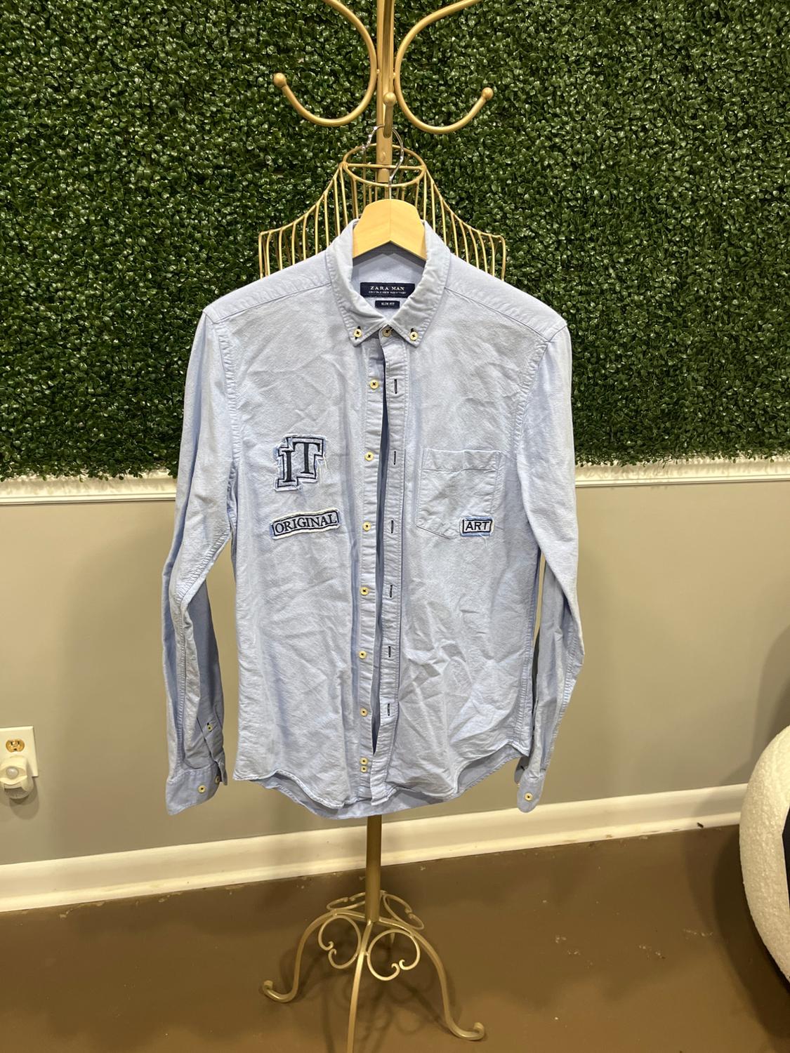 Light blue shirt with "TF", "ORIGINAL", and "ART" patches hanging on a golden rack in front of a green plant background.