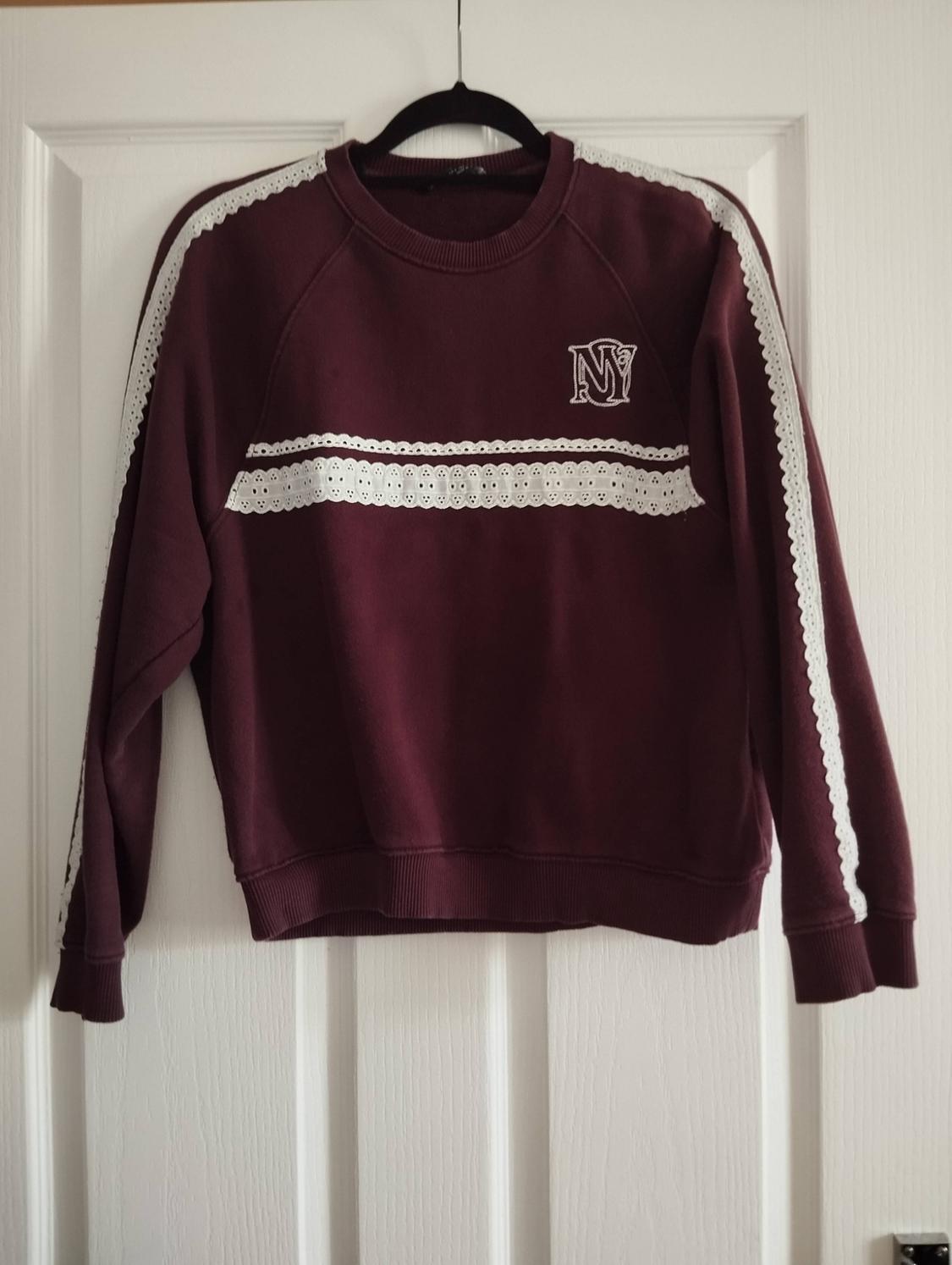 Maroon sweatshirt hanging on a white door. Featuring white lace detailing on the chest and sleeves, and the text 'NY' embroidered.