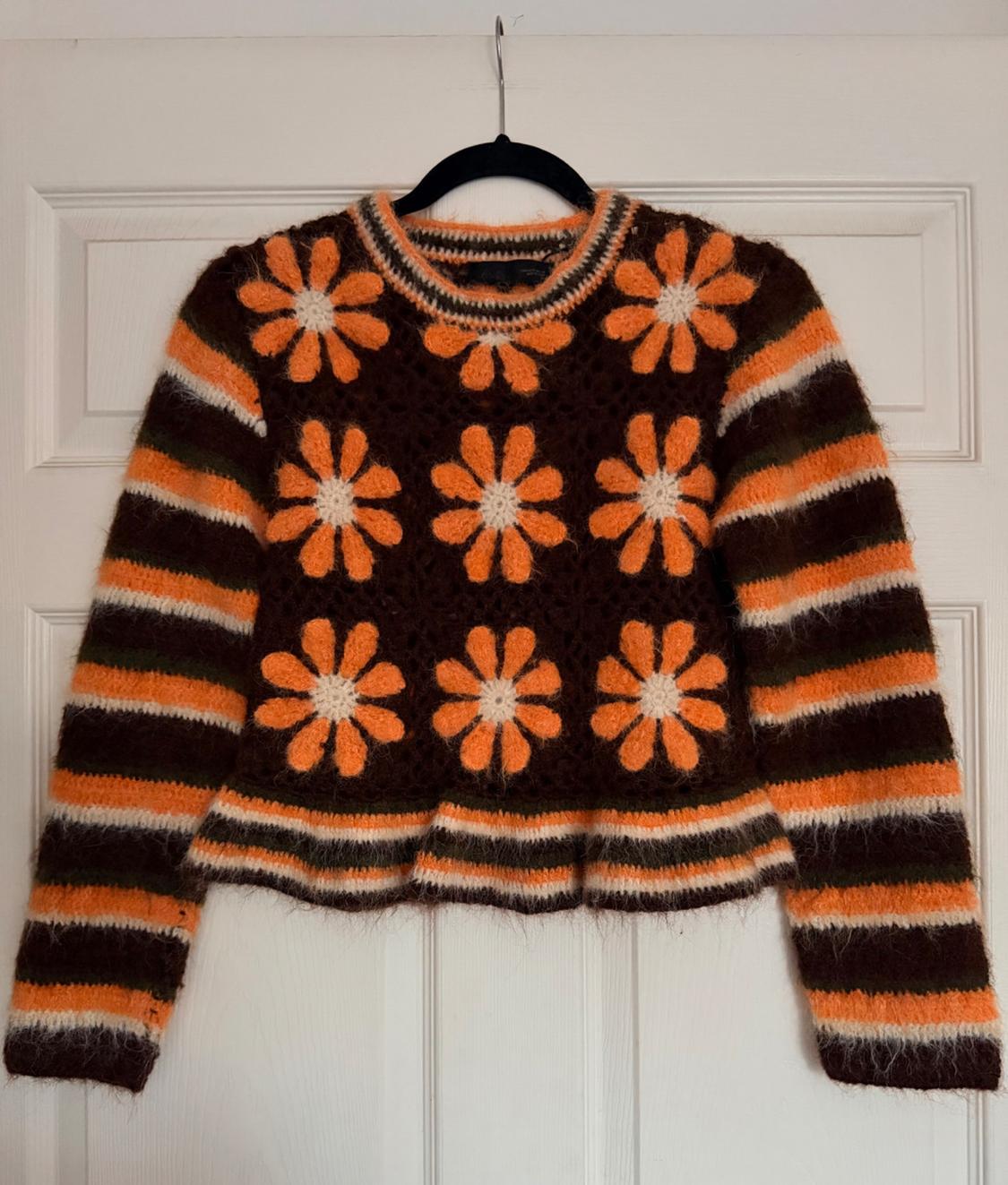 Brown wool sweater with orange and white flowers, and brown, orange, and white striped sleeves, hanging on a white door.
