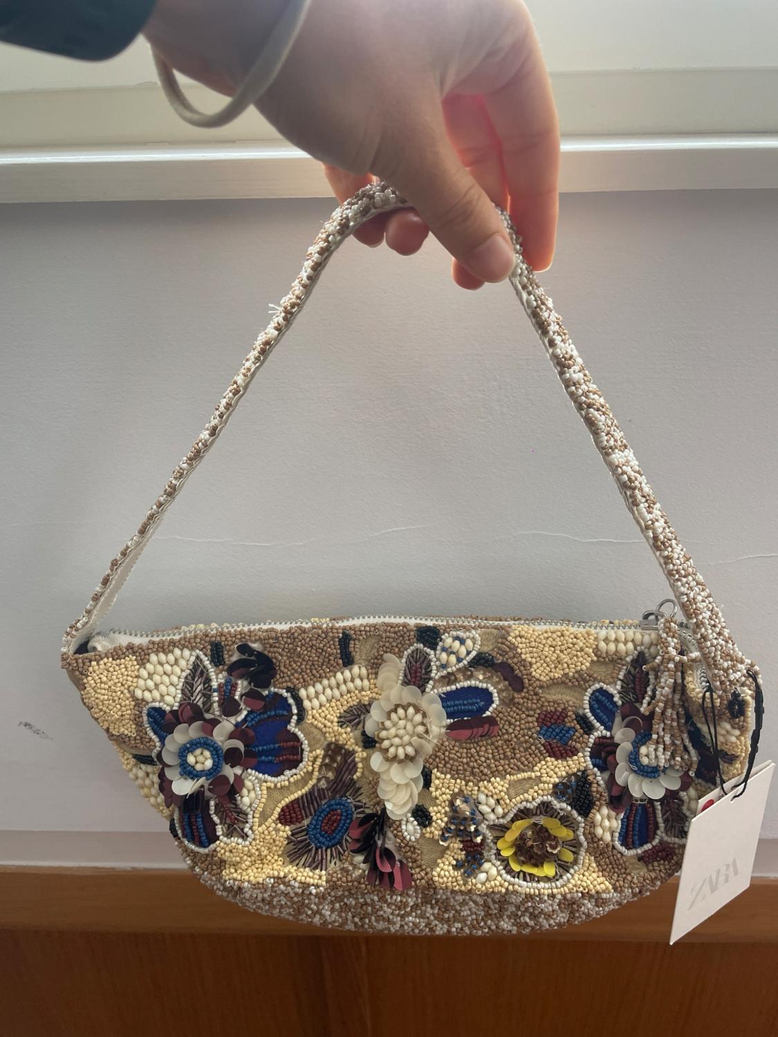 Hand holding a handbag with floral-design beads and sequins. Label bearing the text ZARA. White background.