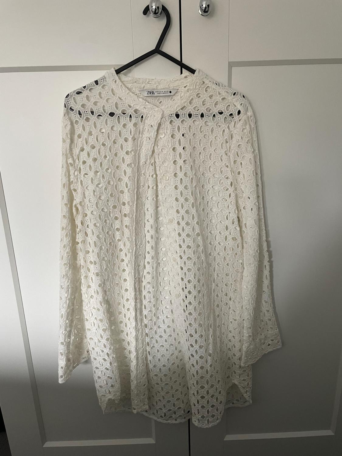 White lace blouse with a circular pattern hanging on a black hanger in front of a white wardrobe. Label visible.