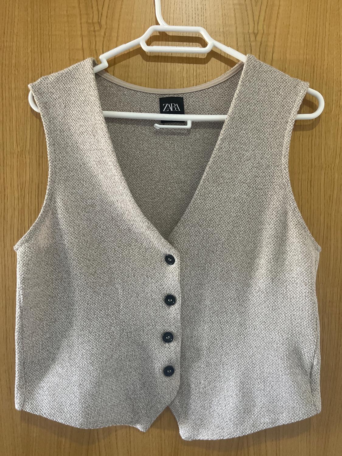 Light grey waistcoat with black buttons hanging on a white hanger over a wooden door. Visible label: ZARA.