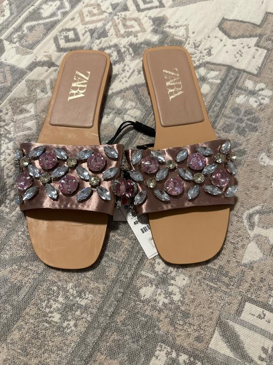 Flat sandals with pink and silver rhinestones on a geometric patterned rug. Visible text: "ZARA".
