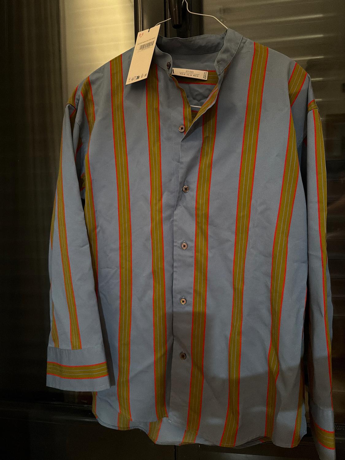Vertical striped shirt in blue, green, and orange, hanging on a hanger with a visible tag, dark background.