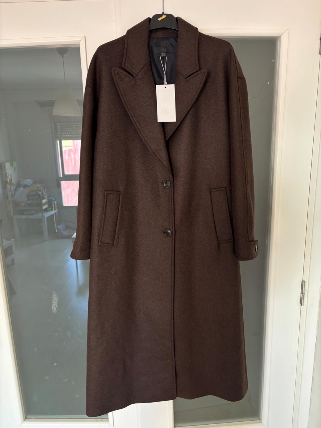 Long brown coat hanging on a hanger in front of a glass door. White label visible on the collar. Indoor setting.