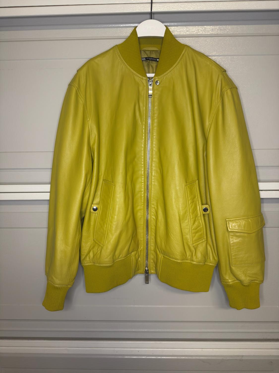 Yellow bomber jacket with a zipper, buttoned pockets, and ribbed trim, hanging on a hanger against a white paneled background.