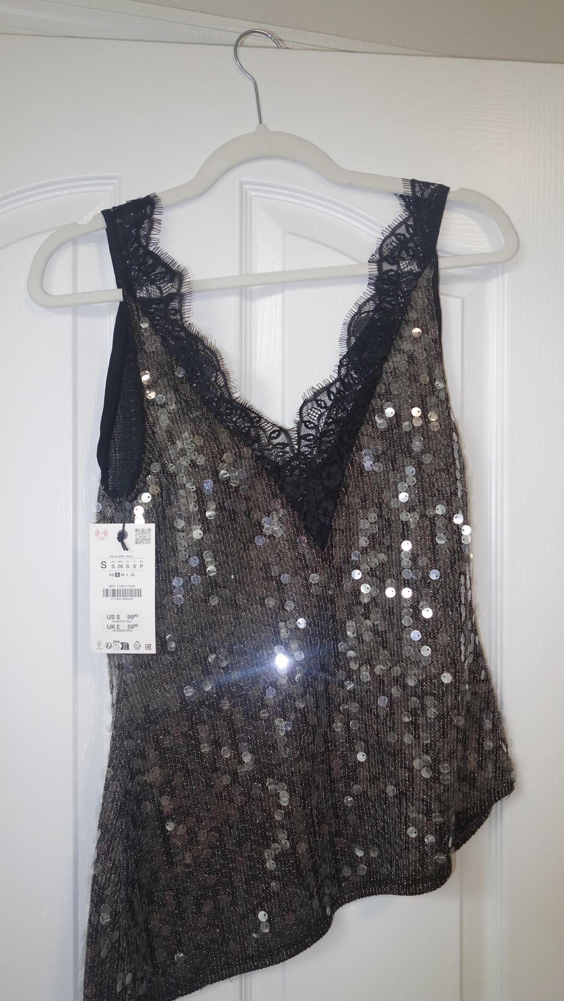Silver sequin top with black lace neckline, hanging on a white hanger. Visible tag with price and size.