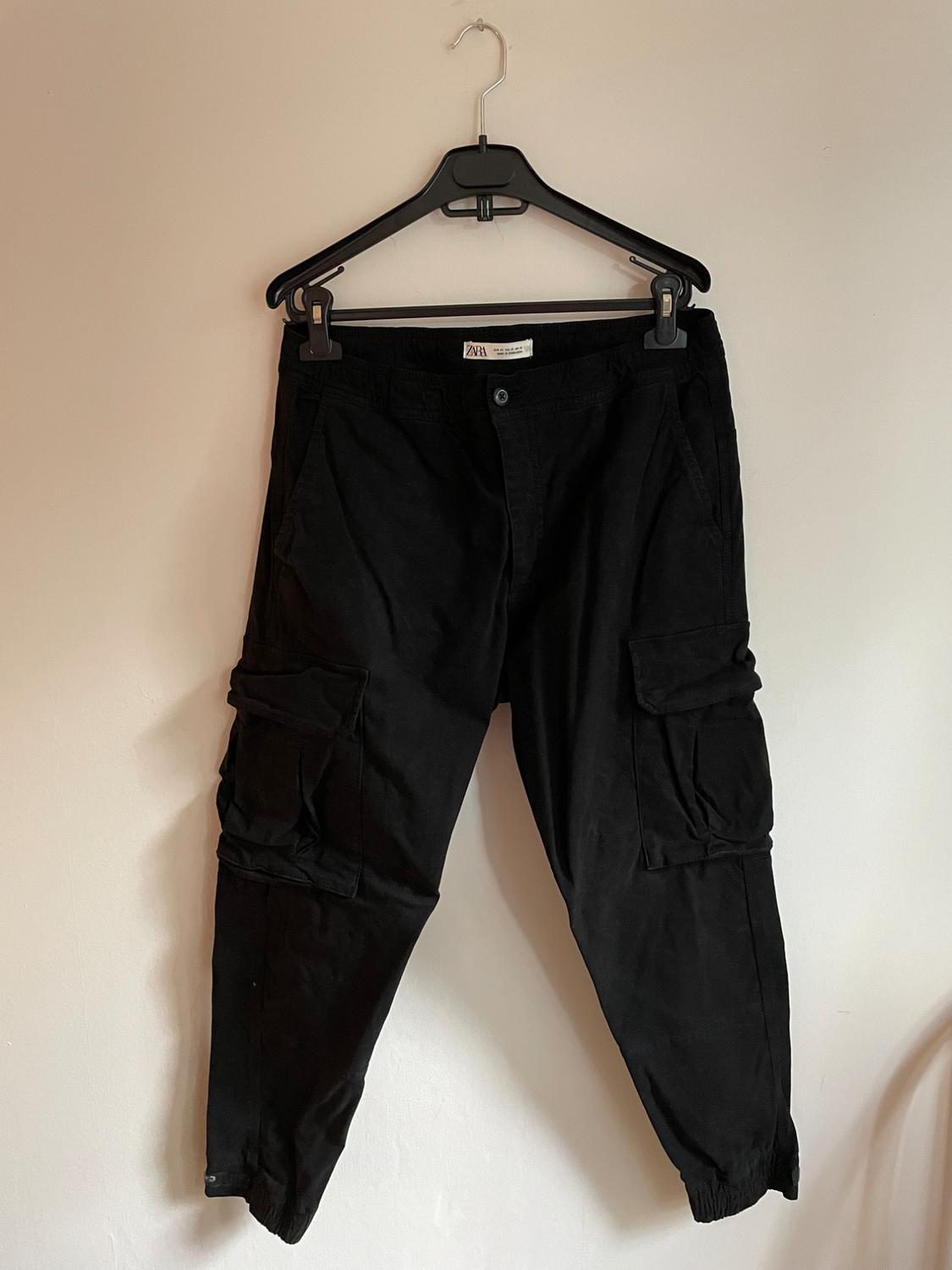 Black cargo trousers hanging on a black hanger against a white wall. Label visible on the waistband.