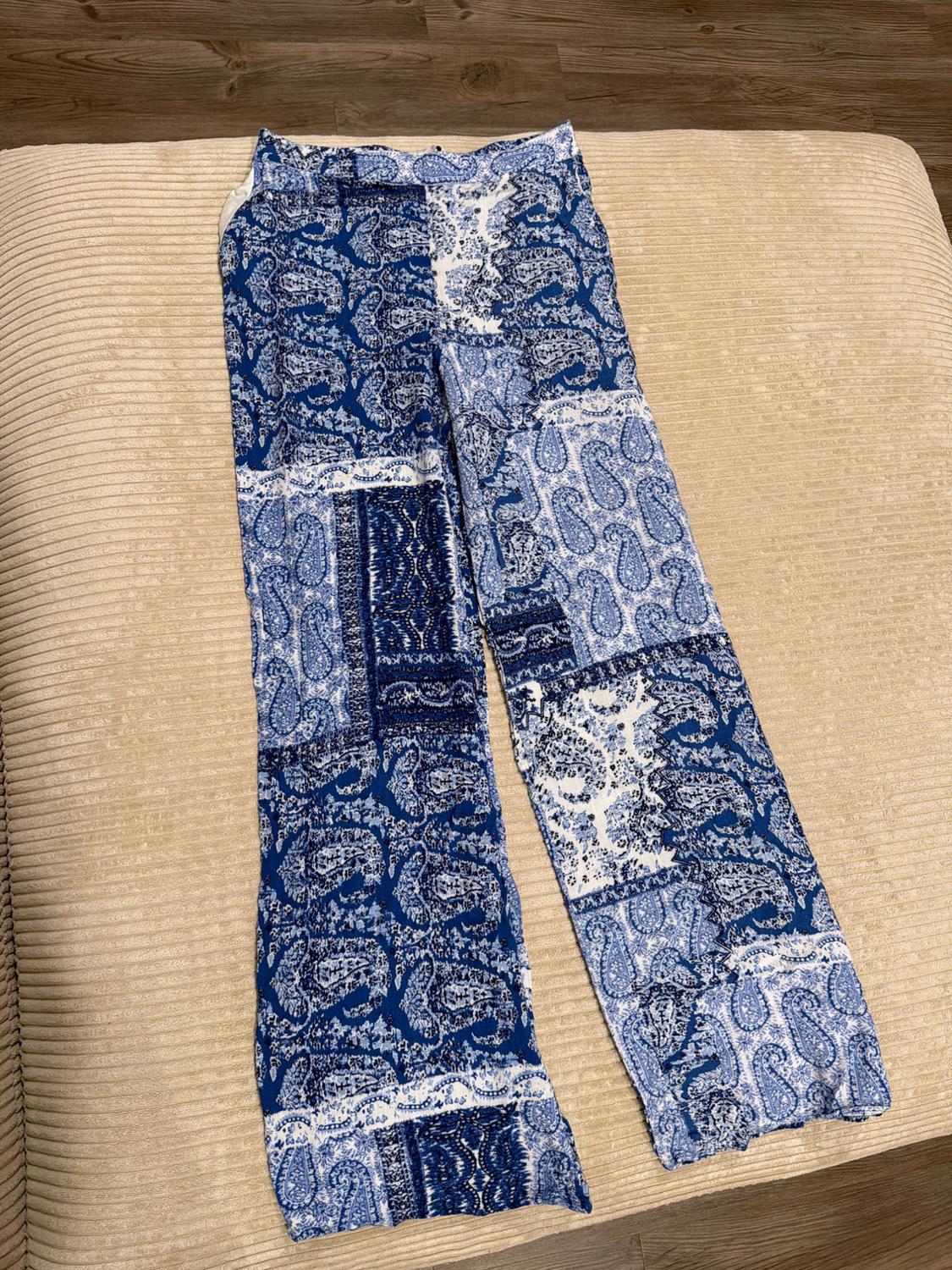 Flowy pants with a blue-and-white paisley patchwork print, on a ribbed beige surface and wooden floor.