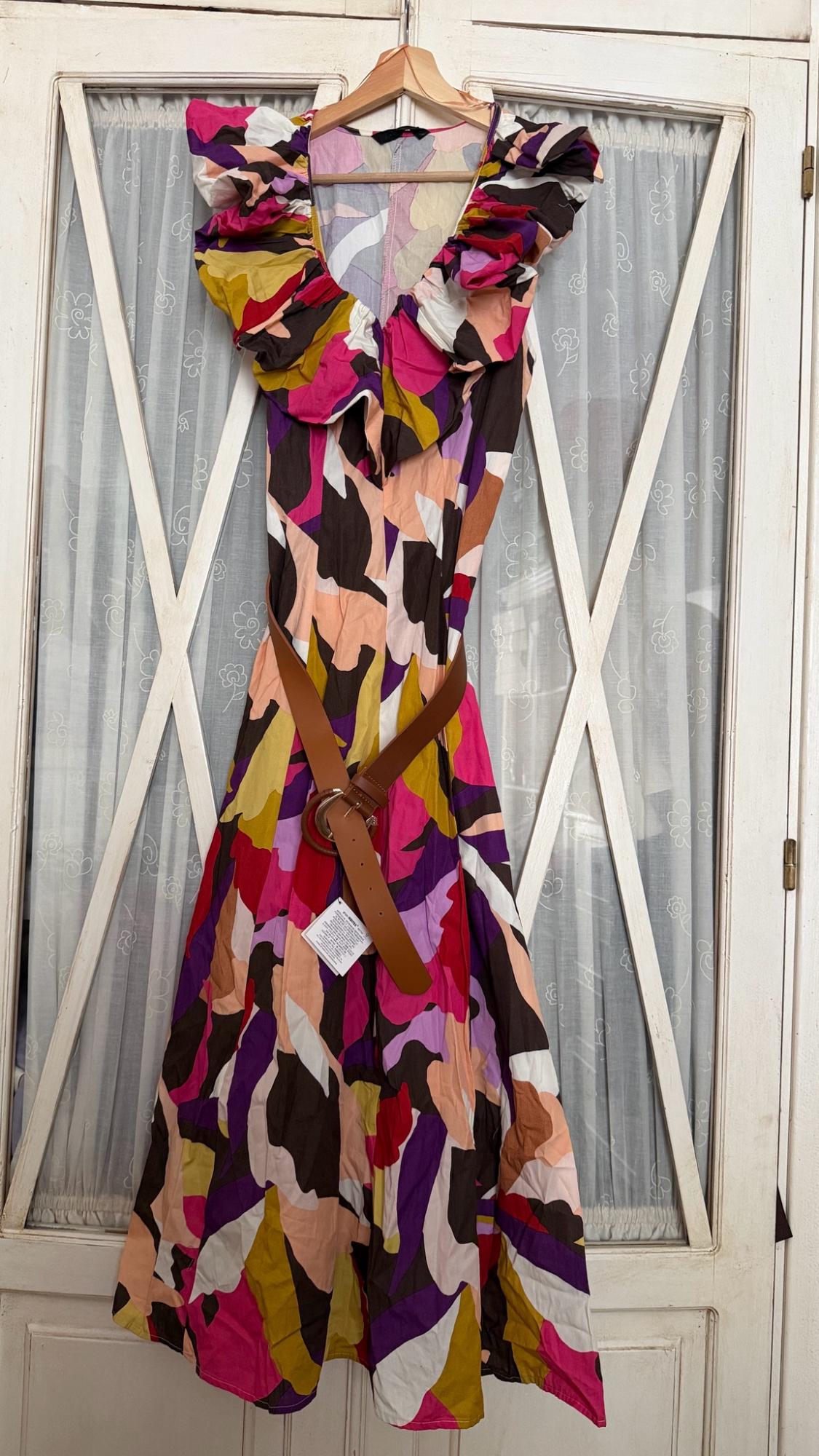 Multicoloured dress with an abstract print, ruffles and a brown belt, hanging on a white door with glass panels and curtains.