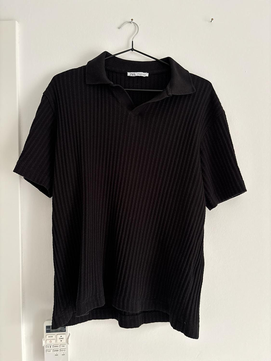 Black ribbed short-sleeved polo shirt on a hanger against a white wall. Legible text: ZARA, MODE, TEMP, SWING, FAN.